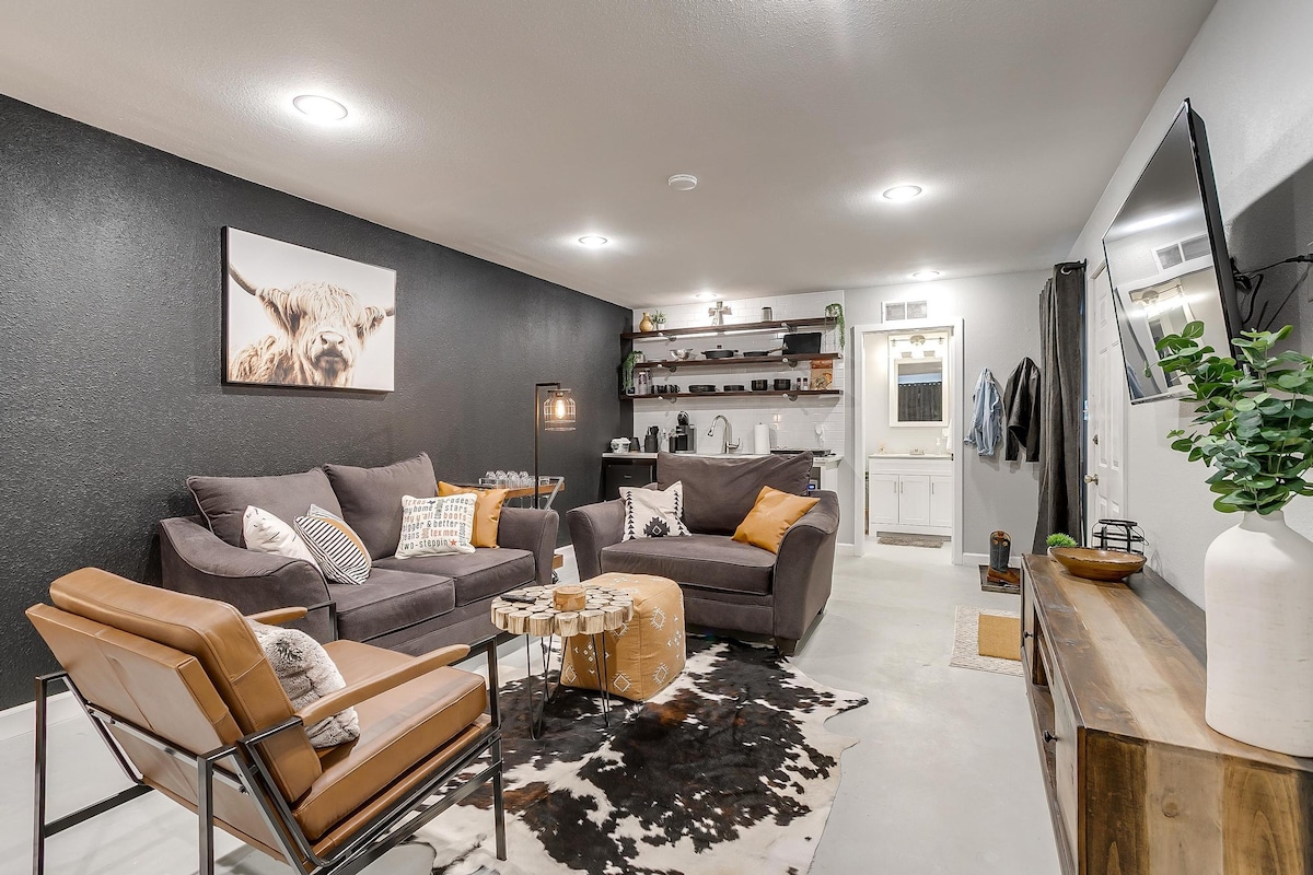 A cozy living area features a dark accent wall and comfortable seating arrangements, including a spacious sofa and leather chair. Decorative touches include a patterned area rug and a wall-mounted TV. Shelves with various items are present, alongside an inviting kitchenette area.