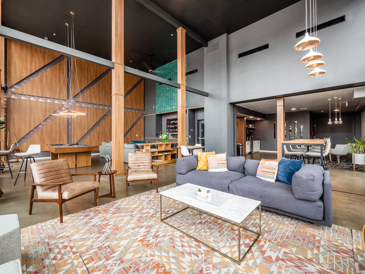 A spacious living area features a sectional sofa with decorative cushions, complemented by two wooden armchairs. A stylish coffee table rests on a patterned rug, with soft lighting provided by pendant fixtures hanging from the high ceiling. The open design includes functional workspaces in the background.