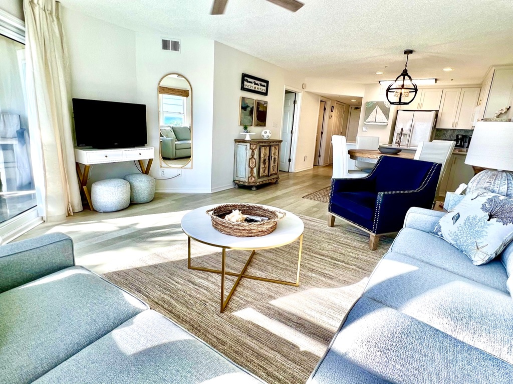 A light-filled living area features a modern blue sofa and a white coffee table with a decorative basket. A stylish accent chair and entertainment console are visible, complementing the open layout that leads to the dining area and kitchen.