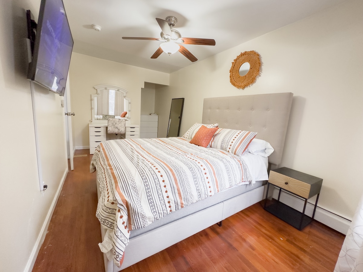 The cozy bedroom features a plush bed with a patterned blanket, complemented by decorative pillows. A wall-mounted smart TV is visible, along with a dresser and mirror in the background. A ceiling fan provides added comfort, enhancing the peaceful ambiance.