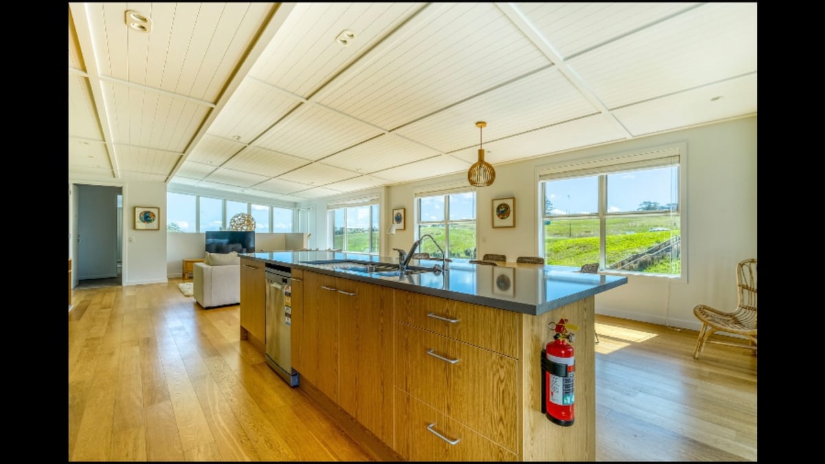 A spacious kitchen features an expansive island with a dark countertop and a stainless steel sink. Large windows allow natural light to fill the space, providing views of the surrounding landscape. Light wood cabinetry complements the room's overall design, while a fire extinguisher is mounted for safety.