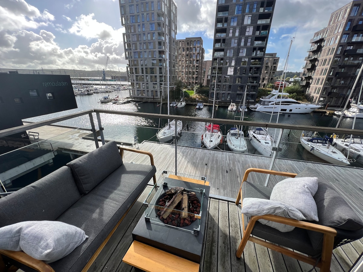 A spacious outdoor seating area features a glass table with a central fire pit, surrounded by modern gray sofas and wooden chairs. A scenic view of the marina with docked boats is visible beyond the glass railing, framed by contemporary apartment buildings.