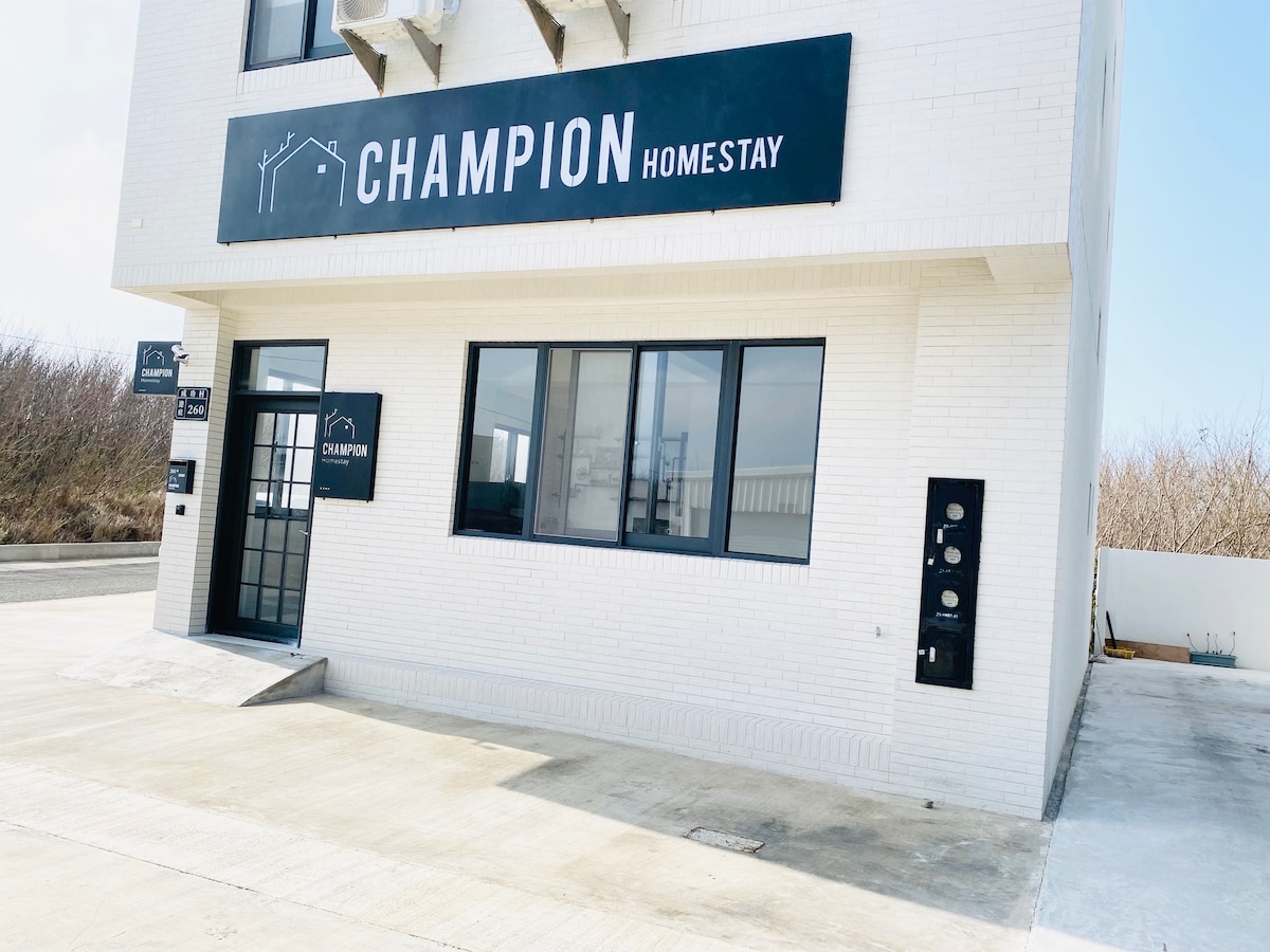 The exterior of Champion Homestay is shown, featuring a modern white building with large windows. The entrance is marked by a black sign displaying the name 'CHAMPION HOMESTAY.' An outdoor area with a concrete surface surrounds the entrance.