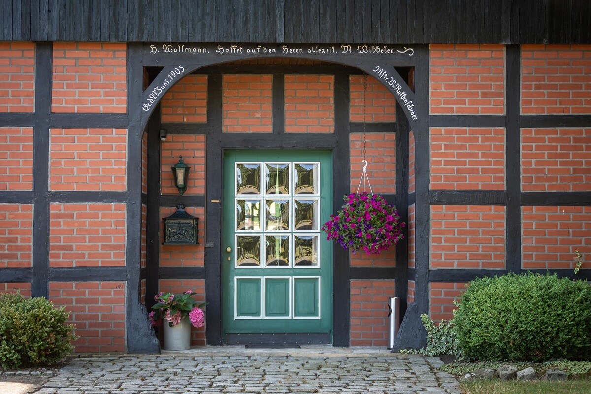 The entrance of a half-timbered house features a large green door framed by decorative brick. A hanging basket of vibrant flowers is placed beside the door, while potted plants add greenery to the stone pathway leading to the entrance.