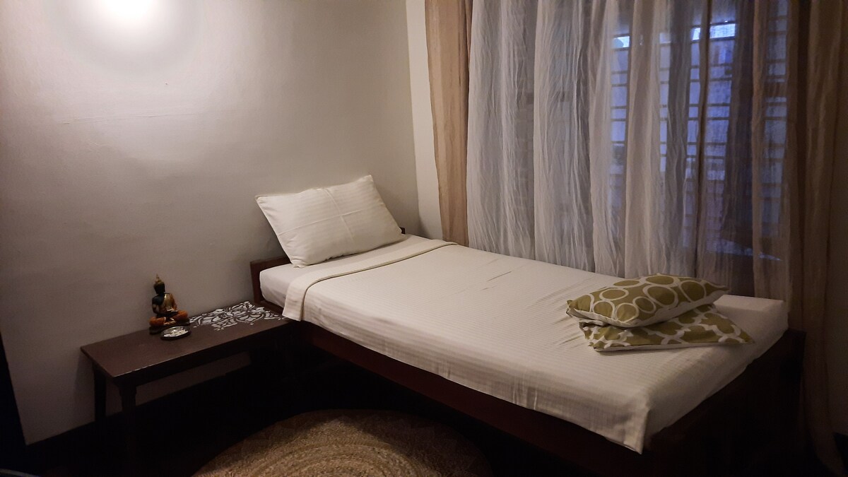 A simple sleeping area is highlighted by a spacious bed adorned with a white cover and decorative pillows. A small wooden table is positioned nearby, and a dim light illuminates the room. Sheer curtains allow gentle light to filter through a nearby window.