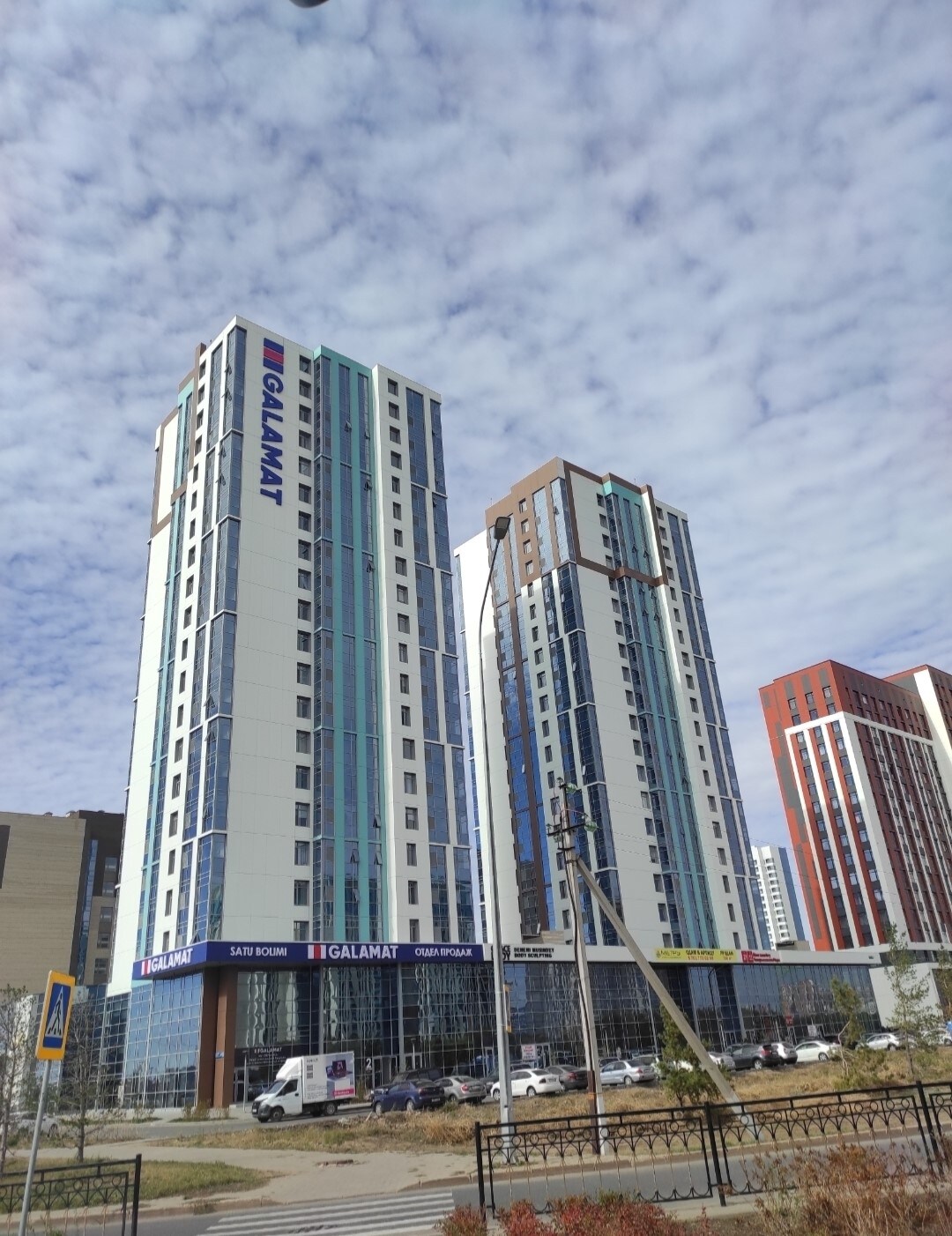 Two modern residential towers are showcased, featuring sleek, glass facades and distinct architectural designs. The Galamat building logo is prominently displayed, reflecting a contemporary urban environment. The surrounding area includes greenery and a mix of paved pathways and vehicles parked nearby.