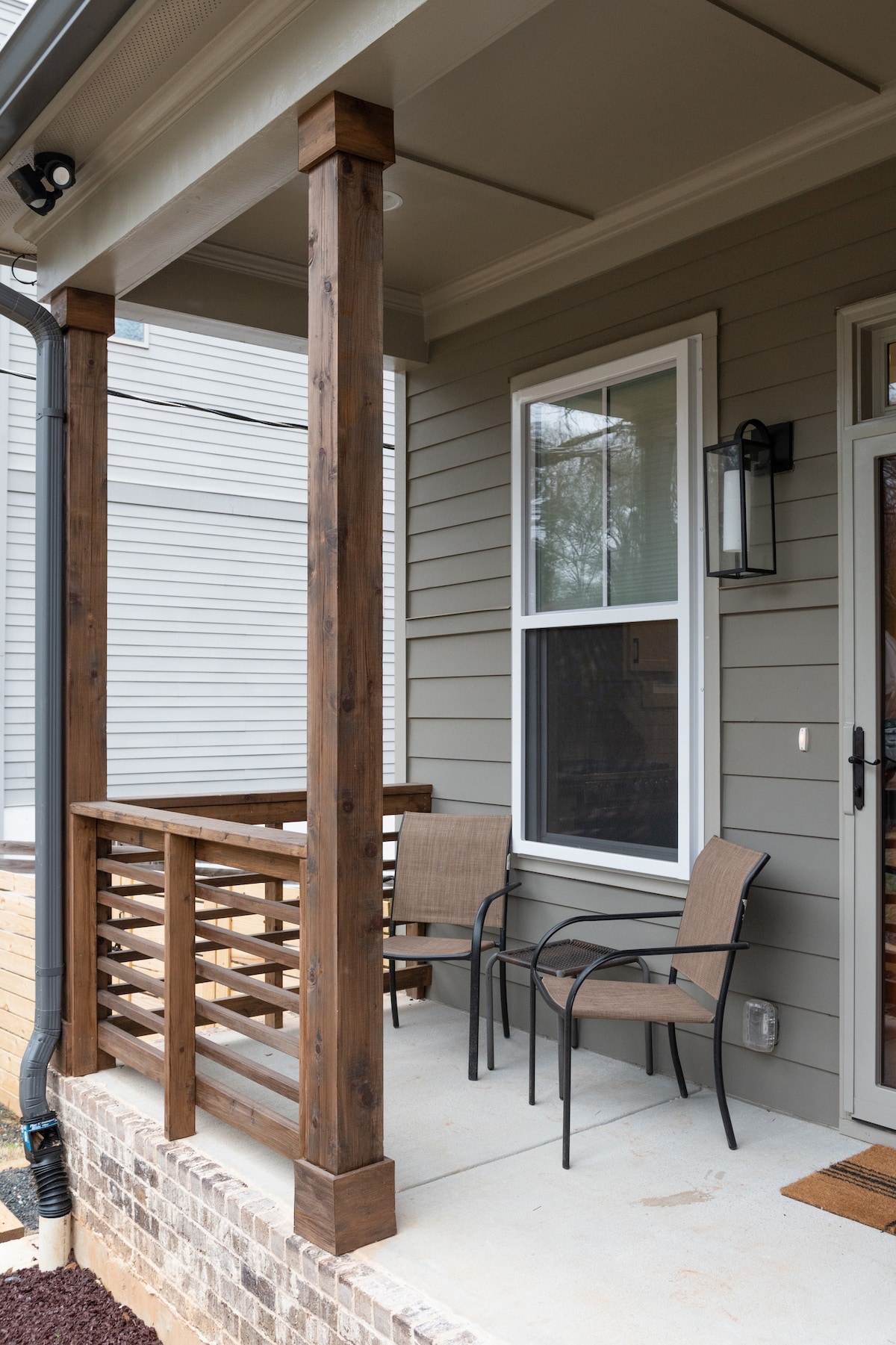A covered front porch features two light brown chairs positioned beside a wooden railing. A small table sits between the chairs, inviting relaxation in this outdoor space. Natural light filters through the nearby window, which is framed by gray siding.