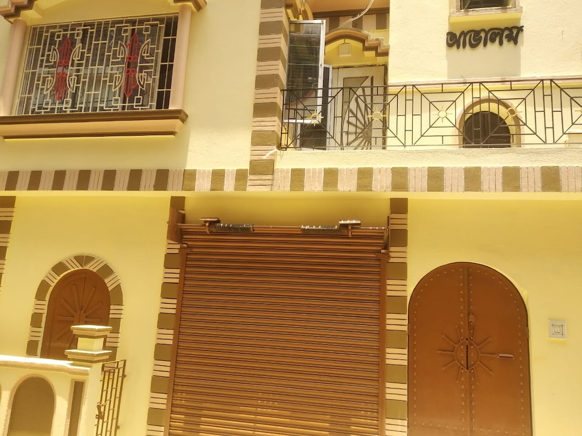The exterior of a two-story building is showcased, featuring decorative elements in a beige-yellow hue. An ornate metal gate is visible, along with a roller shutter that resembles a classic design. A balcony is positioned above, adorned with elegant wrought-iron railings.