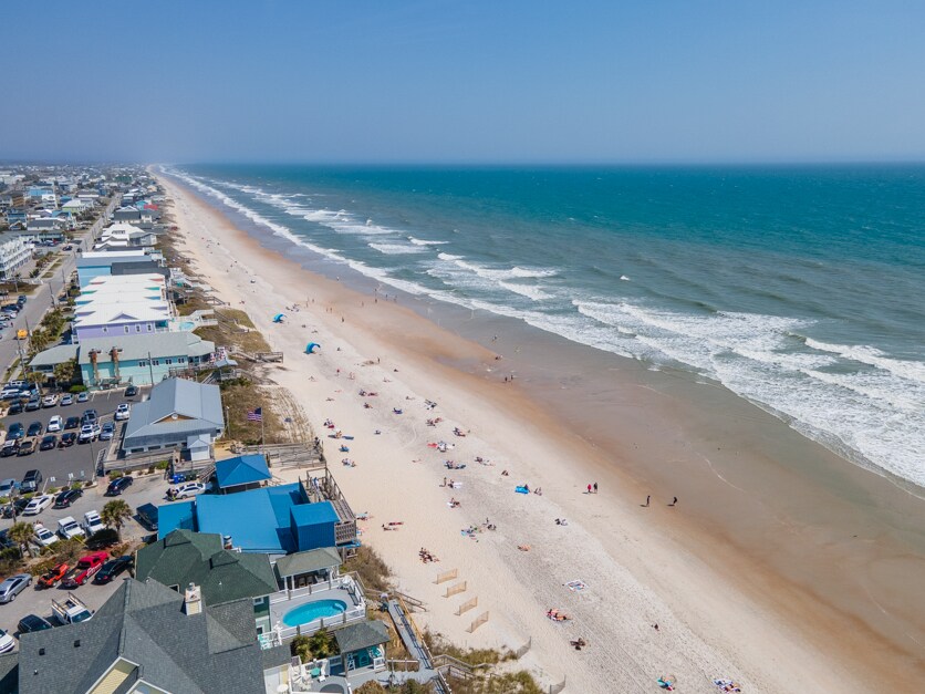 A sweeping aerial view captures the expansive coastline, with soft sandy beaches meeting vibrant blue ocean waves. The scene includes distant beachgoers enjoying the shore, while a collection of buildings line the beach, offering convenience to visitors.