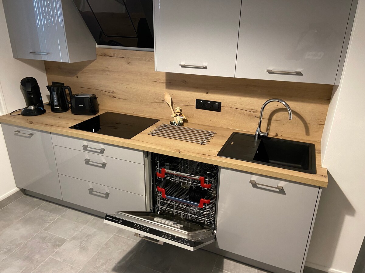 A modern kitchen area features sleek grey cabinetry with wooden countertops. Essential appliances are visible including a coffee maker, microwave, and dishwasher with open drawers. The kitchen sink is positioned under the cabinetry, complemented by a sleek faucet.
