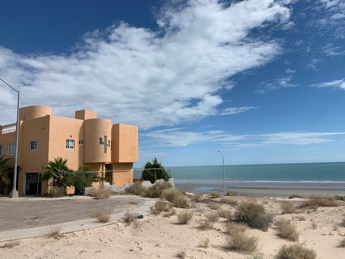 A beachfront property is visible, featuring a unique architectural design with rounded edges. The exterior is painted in warm tones, contrasting with the clear blue sky and turquoise waters. Sandy terrain surrounds the building, enhancing the coastal atmosphere.
