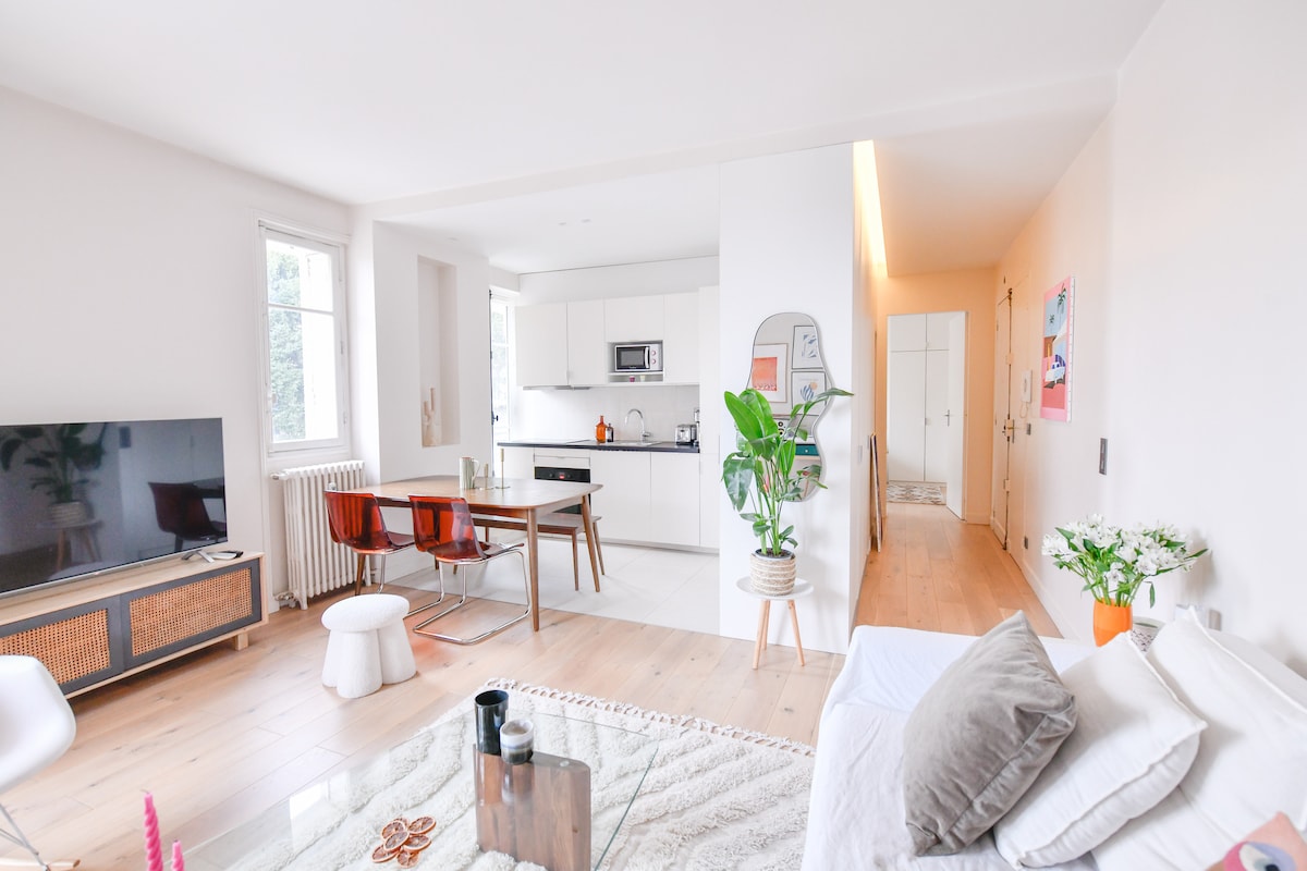 A bright and airy living space is displayed, featuring a comfortable sofa and a large television. A dining table is set beside a fully equipped kitchen. Green plants provide a touch of nature, while light-colored wood flooring enhances the open atmosphere.