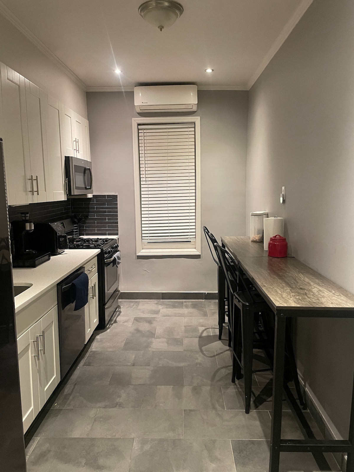 A modern kitchen is equipped with stainless steel appliances, including a gas range and microwave. White cabinets contrast against dark countertops. A small dining area with two high stools is positioned next to the window, which is adorned with horizontal blinds, allowing natural light to enter.