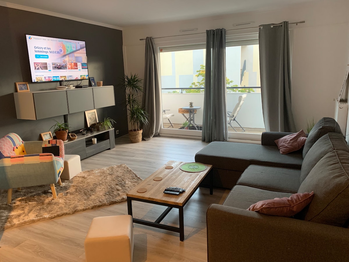 A living area is shown featuring a spacious sectional sofa and a wooden coffee table. A large television is mounted on the wall, accompanied by decorative plants. Natural light enters through sliding glass doors leading to a balcony with an outdoor seating arrangement.
