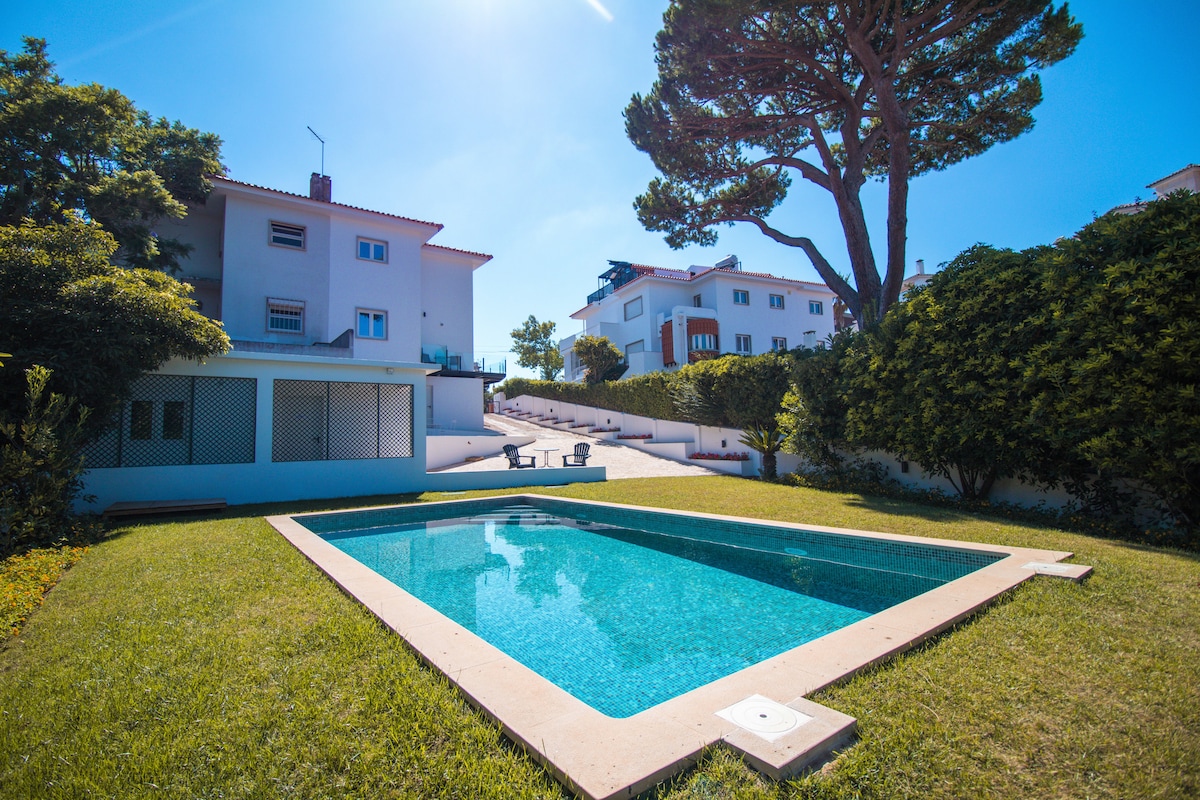 The outdoor area features a rectangular swimming pool surrounded by well-maintained grass. Tall trees and hedges provide a natural backdrop, creating a serene environment. Steps lead from the pool area towards the villa, with lounge chairs positioned nearby for relaxation.