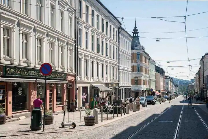 A lively street scene showcases a row of charming buildings with varied architectural styles and shops lining the walkway. Tram tracks are visible, highlighting the convenient public transport. Sunlight bathes the area, enhancing the casual atmosphere with outdoor seating at local establishments.