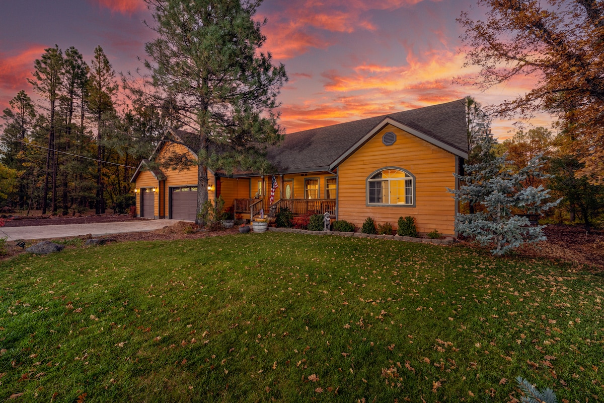 The exterior of a warm wooden house is framed by tall pine trees and a manicured lawn. A two-car garage is visible on the left. The sky is a blend of vibrant colors as the sun sets, adding a serene atmosphere to the home's welcoming front porch.