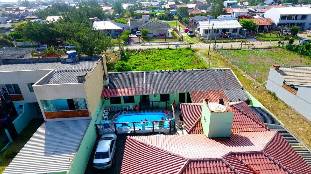 An aerial view captures two adjacent houses, surrounding a central swimming pool. A mix of rooftops is visible, with a vehicle parked nearby. Lush green space and neighboring residences create a tranquil atmosphere. The area is well-defined, showing the layout of the property.
