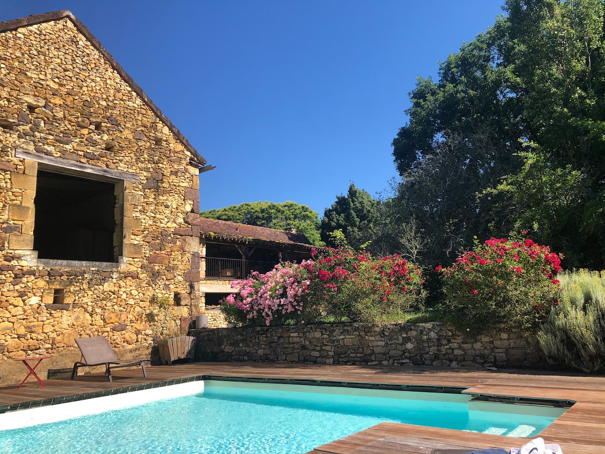 A rustic stone house is featured beside a private swimming pool, surrounded by blooming pink flowers and lush greenery. Sunlight reflects off the clear water, providing a serene atmosphere. A wooden deck borders the pool, offering space for relaxation under the blue sky.