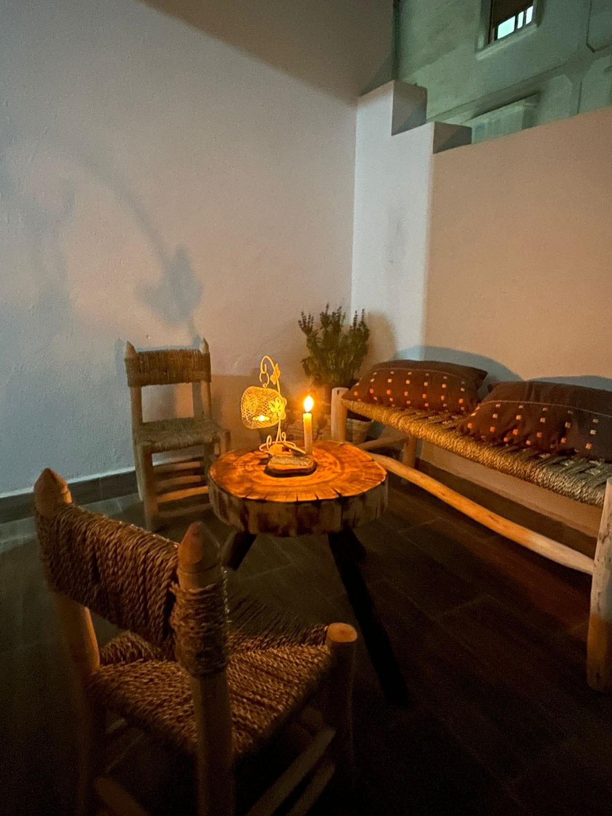 An outdoor seating area is illuminated by a candle's soft glow. The space features two rustic chairs and a round wooden table, complemented by a bench adorned with patterned cushions. Natural elements and textures create a cozy ambiance, suitable for relaxation.