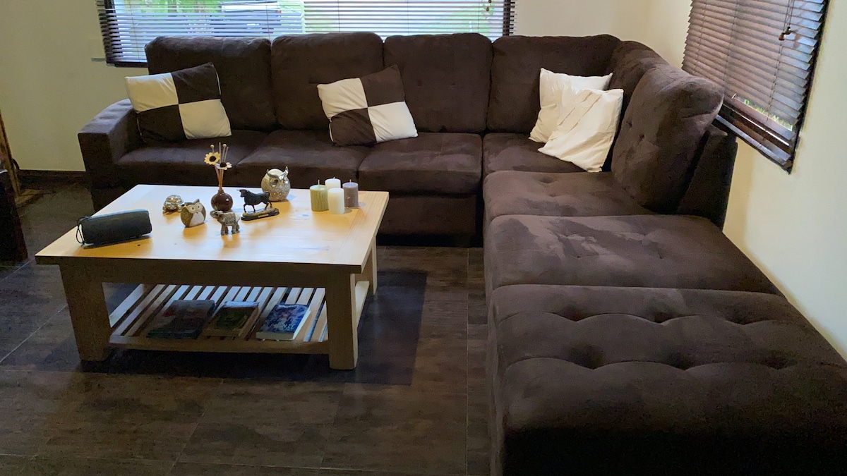 A spacious sectional couch is situated in a cozy living area, adorned with contrasting black and white cushions. A wooden coffee table rests in front, displaying decorative items and candles. Light filters through the window blinds, softly illuminating the room.