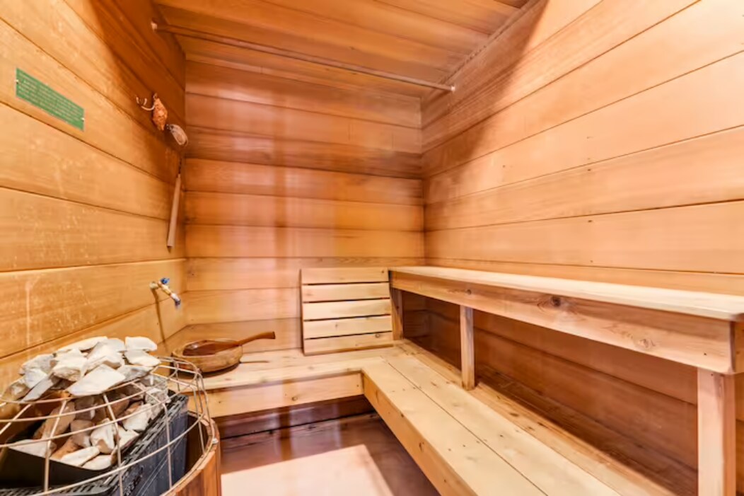 A wooden sauna interior features warm-toned paneling and multiple benches along the walls. A stone heater is visible, accompanied by a wooden ladle. Natural light illuminates the serene space, promoting a relaxing atmosphere.