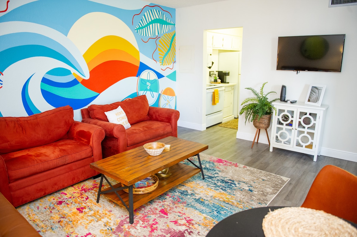 A colorful living area features two vibrant orange sofas arranged around a wooden coffee table. A large mural of waves in bright colors extends across one wall, while a kitchen area is visible in the background. A patterned rug adds texture to the space.