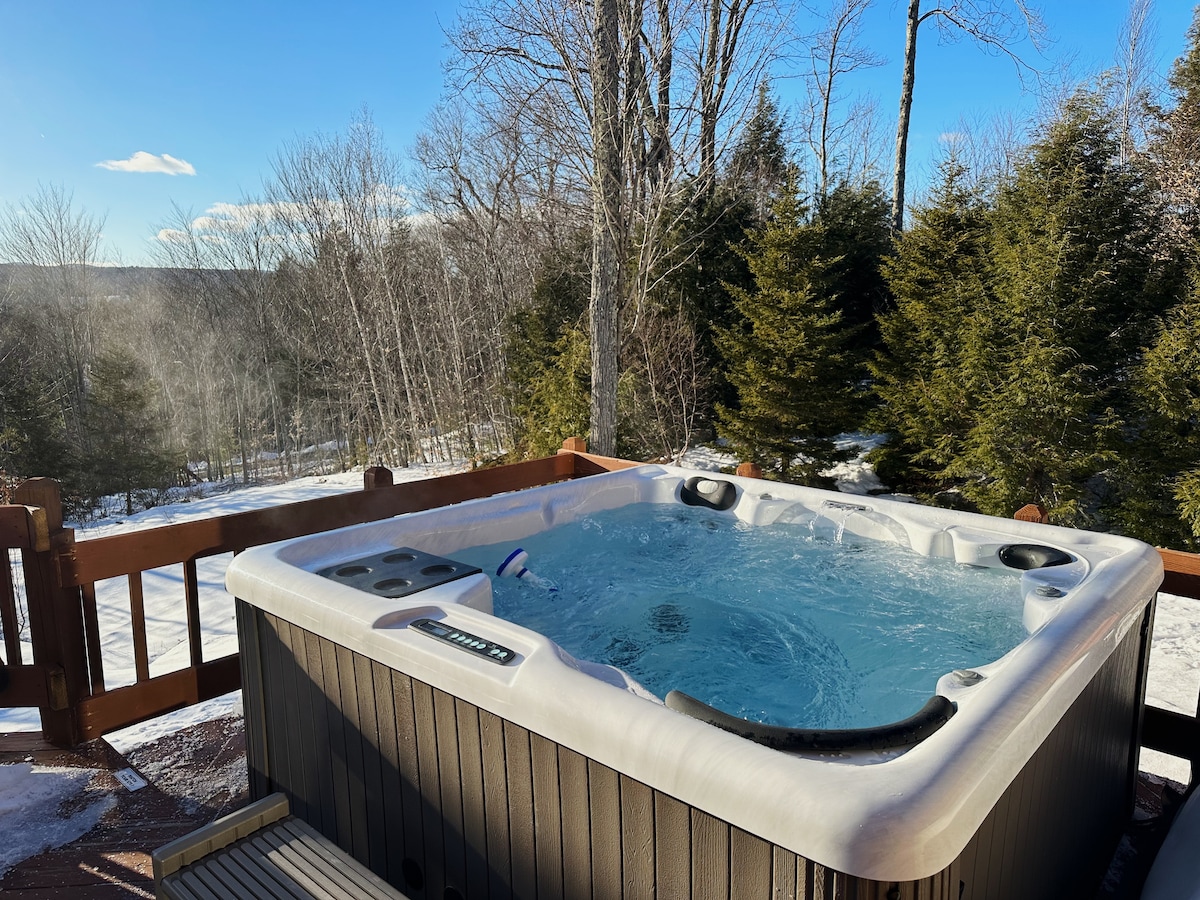A full-size hot tub sits on a wooden deck, with steam rising from the water. Surrounding pine trees are partially covered by snow, offering a serene view of the winter landscape and distant hills.