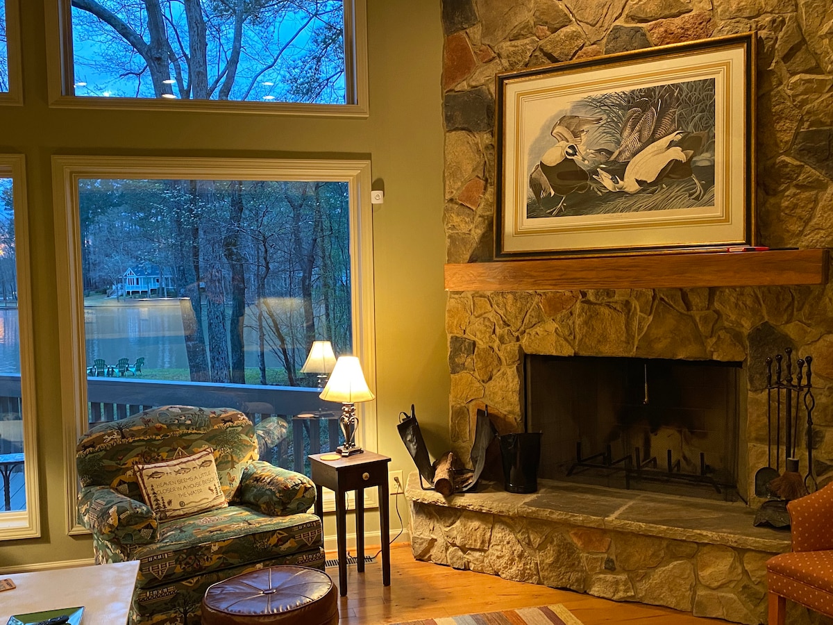 A cozy living area features a natural stone fireplace with a framed artwork above it. A patterned armchair and a small lamp provide comfort, while large windows reveal views of the surrounding nature and evening sky.