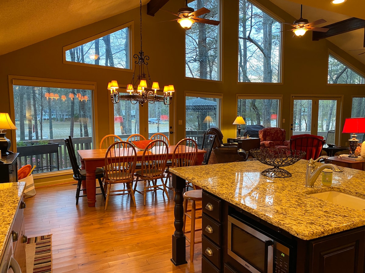A spacious dining area is showcased with a long wooden table surrounded by chairs, located near large windows that offer views of the outdoors. Soft lighting from various lamps complements the warm wood tones of the flooring and furnishings.