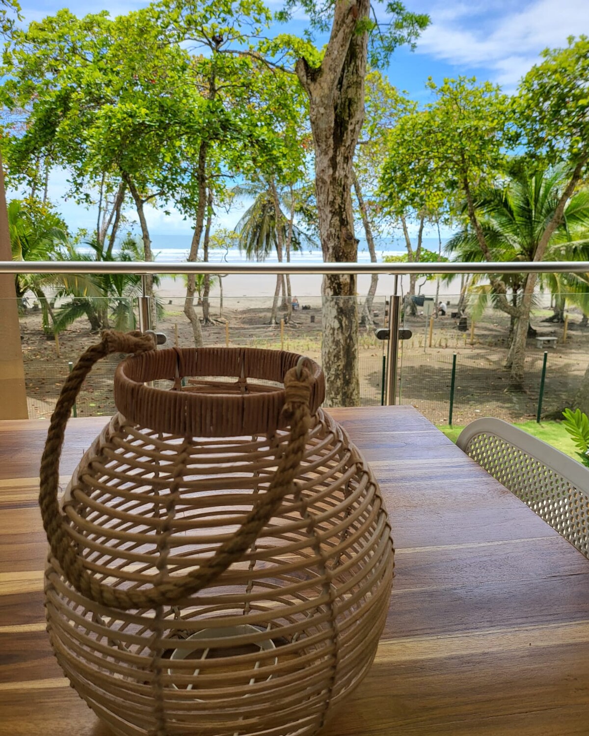 A woven lantern is placed on a wooden table, providing a natural touch. In the background, trees with lush green leaves frame a view of the sandy beach and the ocean, enhancing the serene coastal environment.