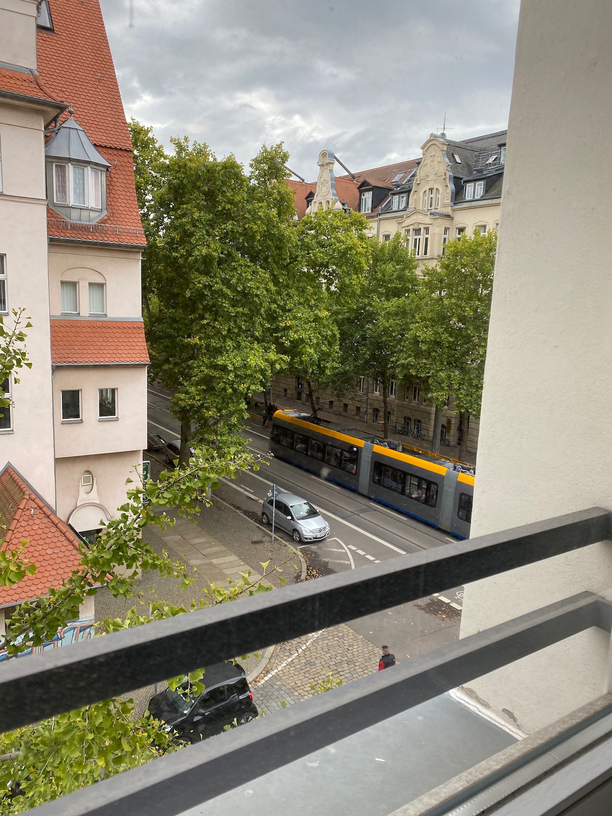 A view from the balcony showcases tree-lined streets and a tram passing below. The architecture features a mixture of traditional and modern buildings, with red rooftops and intricate facades visible in the background. A quiet ambiance is present, with cars parked along the road.