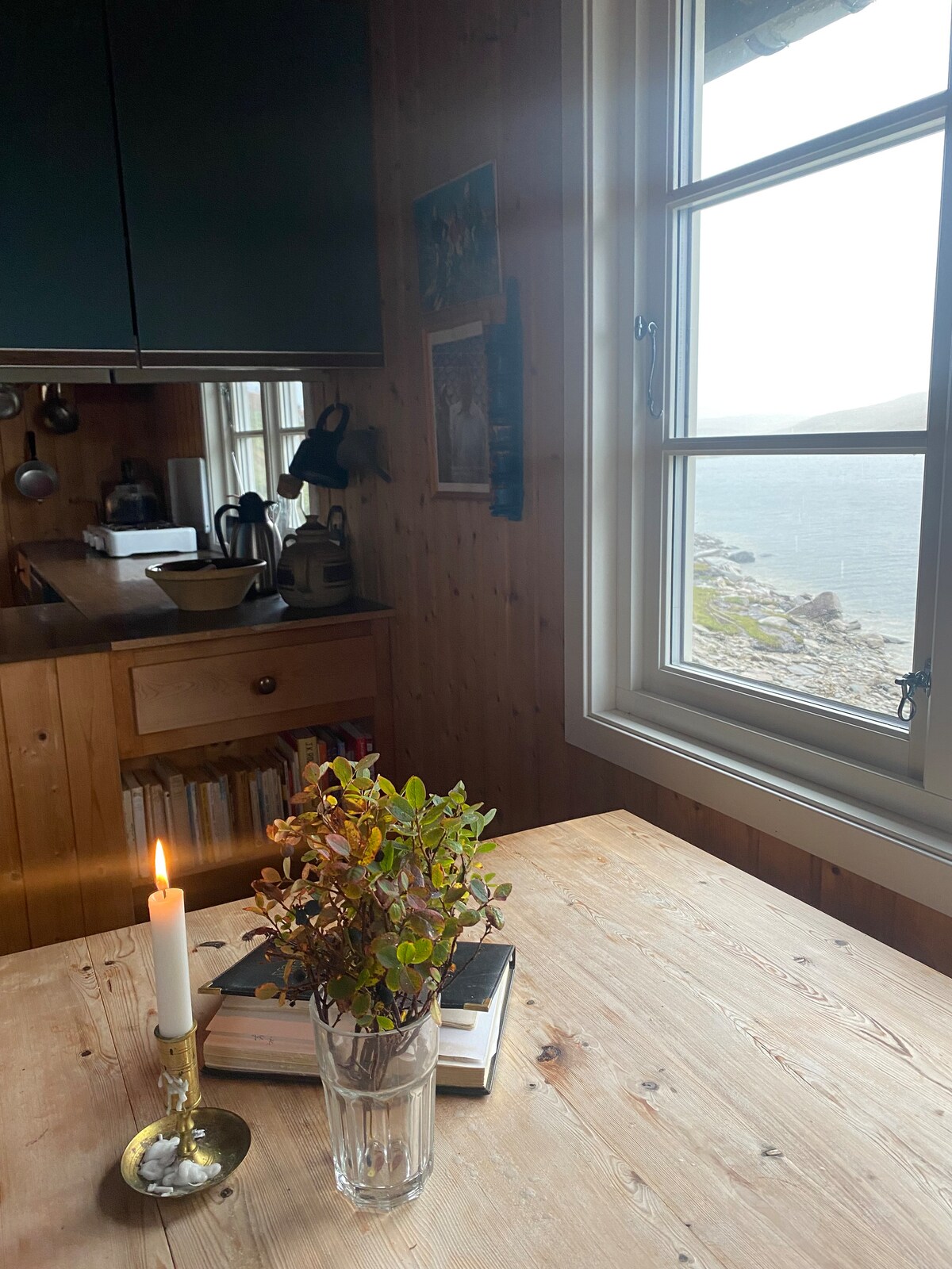 A wooden dining table is seen near a window overlooking a tranquil water view. A lit candle and a small vase with greenery are placed on the table. The kitchen area is visible in the background, featuring wooden cabinetry and kitchenware.