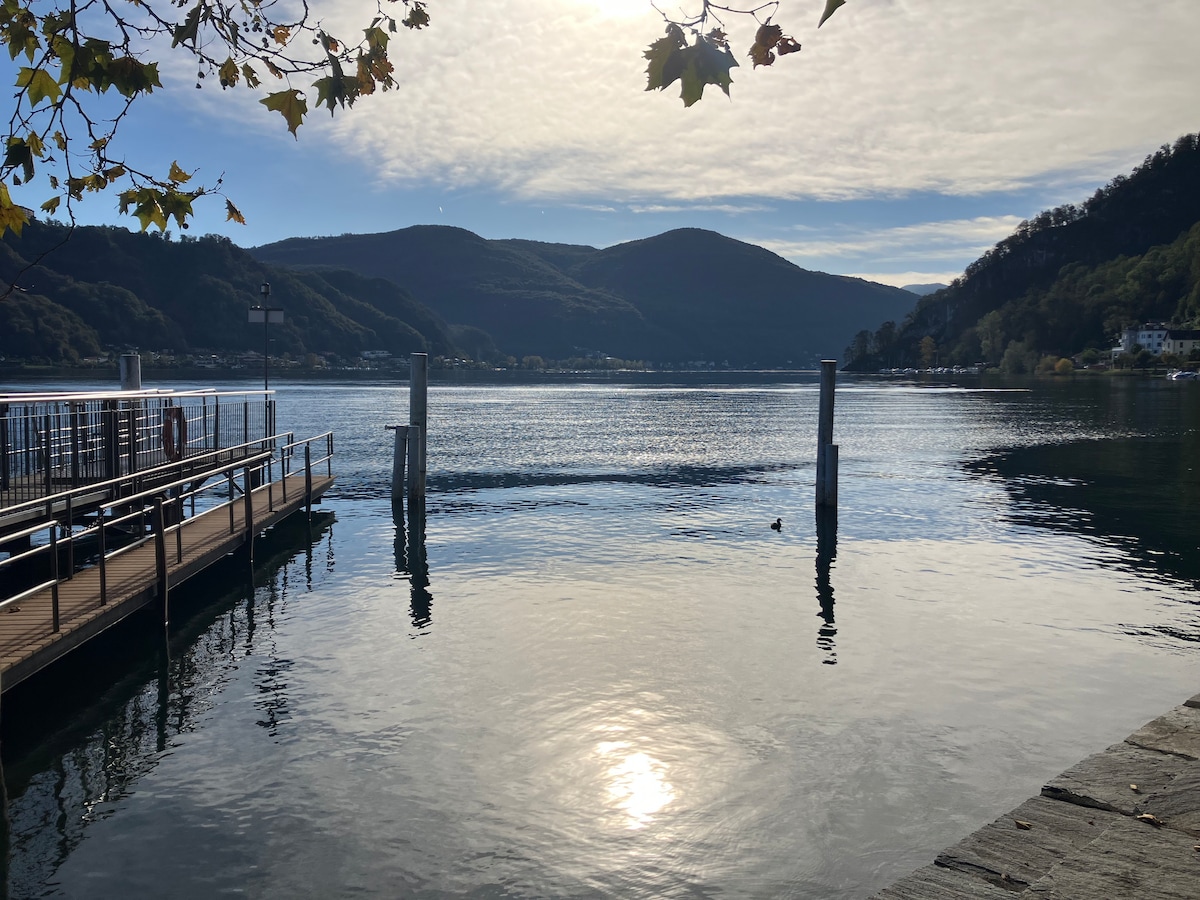 A serene lake scene is depicted, with calm waters reflecting the surrounding hills and clouds. A wooden pier extends into the lake, supported by concrete posts. The area is framed by trees, creating a natural and peaceful atmosphere.