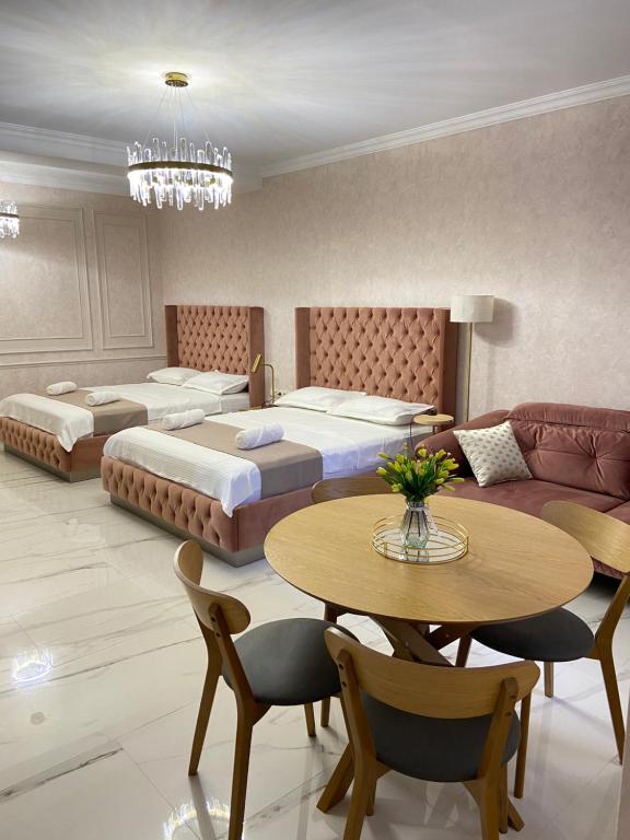 A well-appointed guest room features two spacious beds with tufted headboards. A round wooden table with six chairs is positioned nearby, complemented by a cozy seating area with a sofa. Elegant light fixtures hang from the ceiling, enhancing the room's modern design.