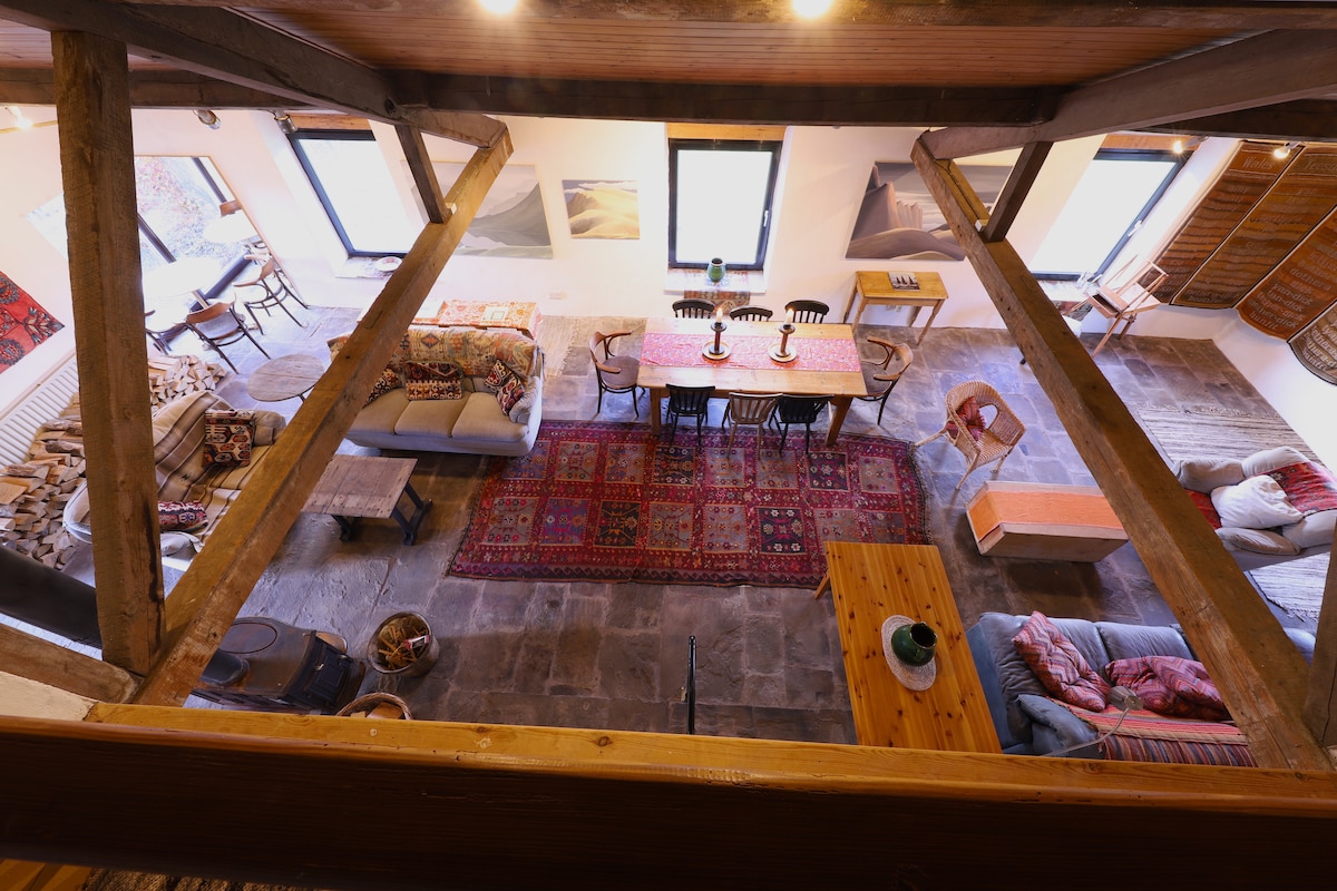 An expansive open plan living area is visible from an elevated perspective, showcasing a blend of seating options, including sofas and chairs. A long dining table centrally features, surrounded by various artworks on the walls. Large windows offer ample natural light, accentuating the stone flooring.
