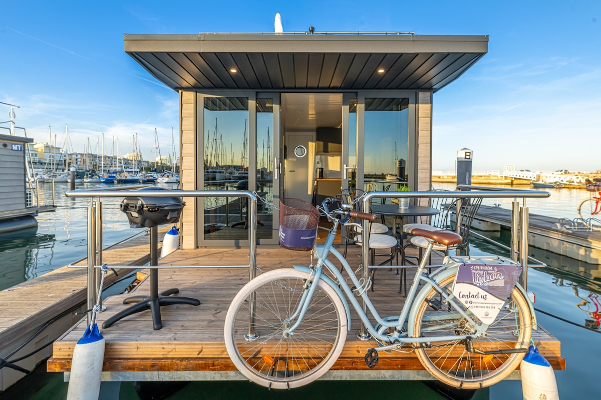 The exterior of a modern homeboat is displayed, featuring a wooden deck extending over the water. A bicycle is parked beside a grill, while large windows allow for light to enter the living space inside. The marina's boats are visible in the background.