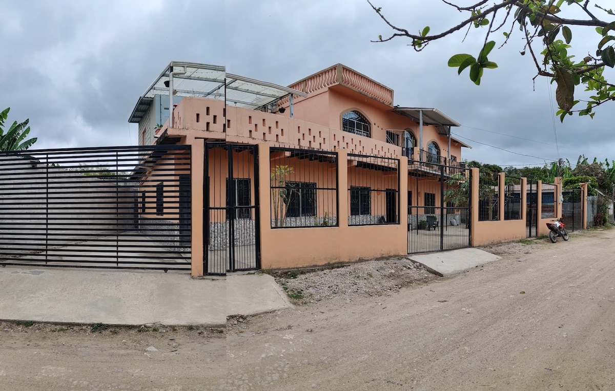The exterior of a large, peach-colored two-story home is shown, featuring a gated entry and a spacious façade. The front is surrounded by a simple metal fence with plants bordering the area. A stone pathway leads up to the entrance.