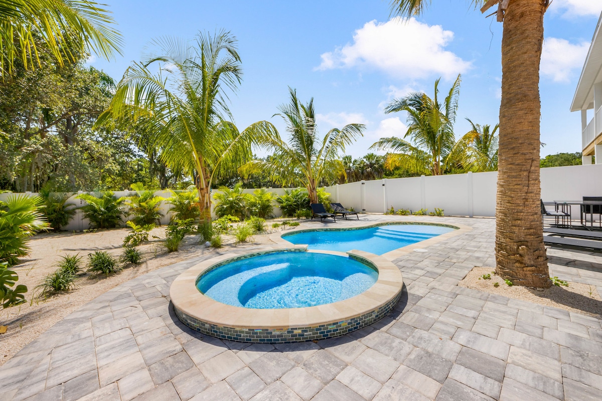 A refreshing backyard features a circular hot tub adjacent to a spacious pool, surrounded by lush palm trees and tropical plants. The area is paved with light gray stone tiles, providing a clean and inviting space for relaxation and enjoyment.