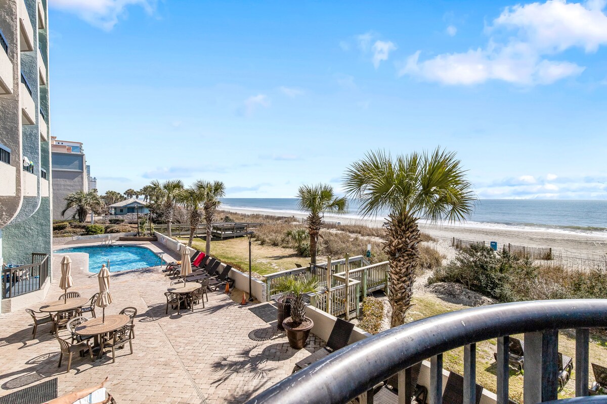 A view from the balcony reveals a large outdoor pool surrounded by lounge chairs and palm trees. Direct ocean access is visible, along with a sandy beach that stretches into the distance under a clear blue sky.