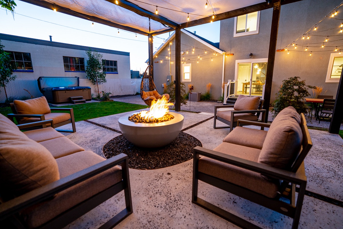 An inviting backyard lounge is featured, showcasing a gas fire pit surrounded by comfortable seating. String lights hang above, enhancing the ambiance. In the background, a hot tub and trimmed greenery are visible, contributing to a cozy outdoor retreat.