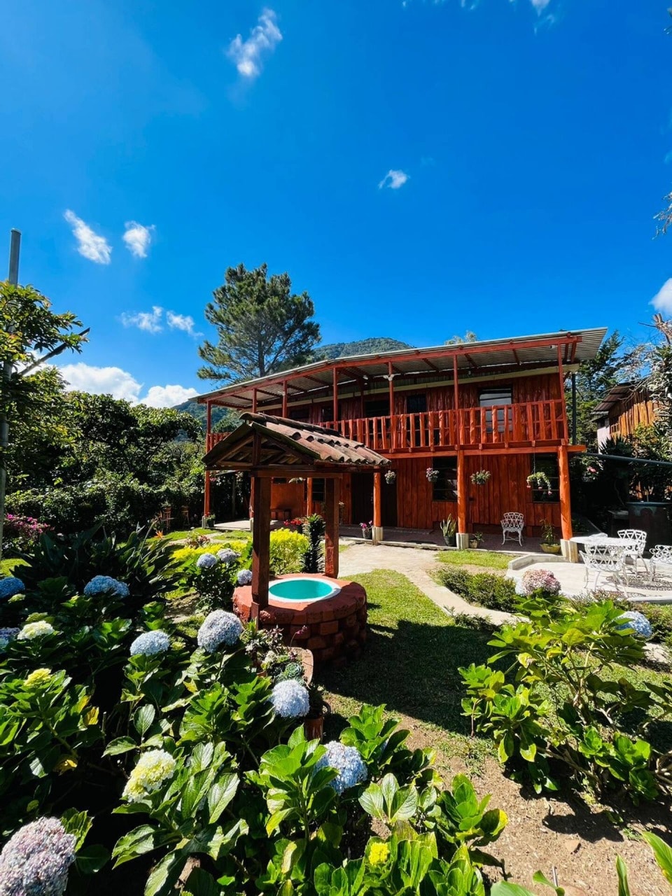 The wooden cabin stands surrounded by vibrant gardens featuring blooming flowers and lush greenery. A small fountain is situated nearby, adding to the tranquil setting. Clear blue skies provide a bright backdrop, enhancing the cabin's inviting exterior with a second floor and balcony.