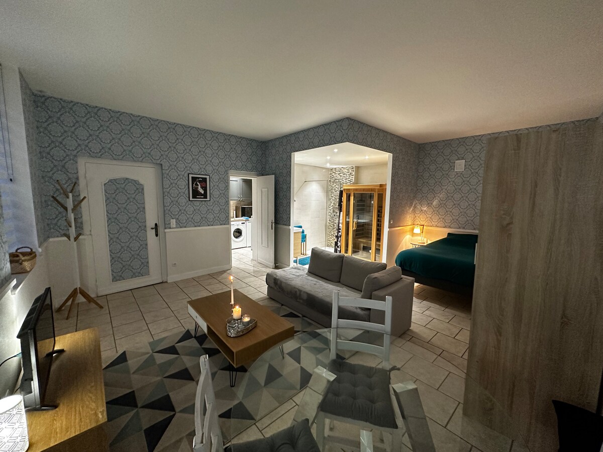 A spacious living area features a neutral-toned sofa and a wooden coffee table with a candle. The walls exhibit a subtle patterned wallpaper. A well-defined sleeping area is visible, showcasing a comfortable bed. The open layout connects to a bathroom area with a sauna in the background.