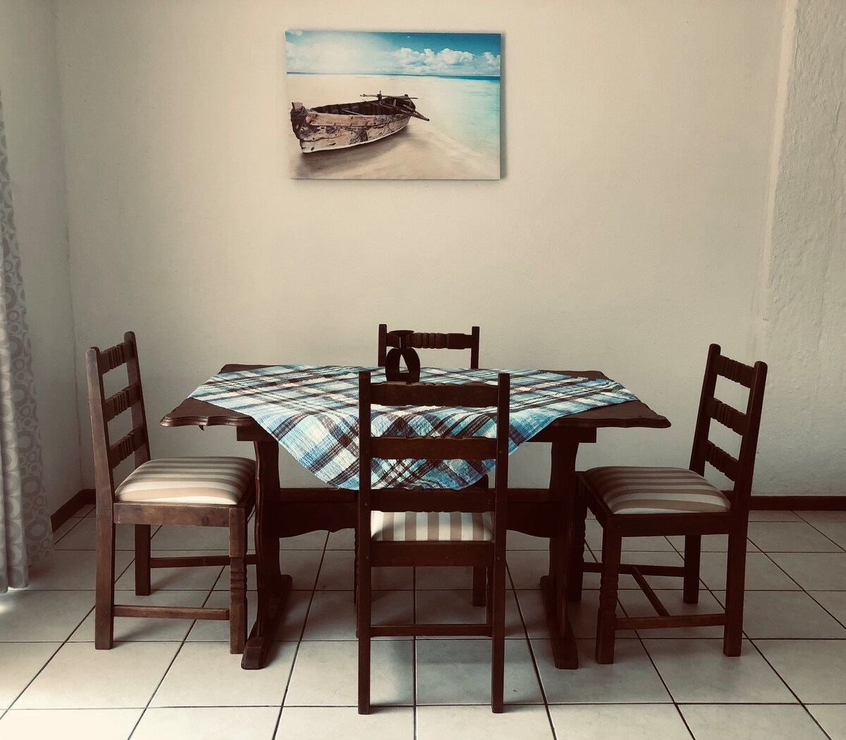 A dining area features a wooden table surrounded by four chairs, two with striped upholstery. A colorful table runner adds a pop of color, while a coastal-themed artwork of a boat rests on the wall above.