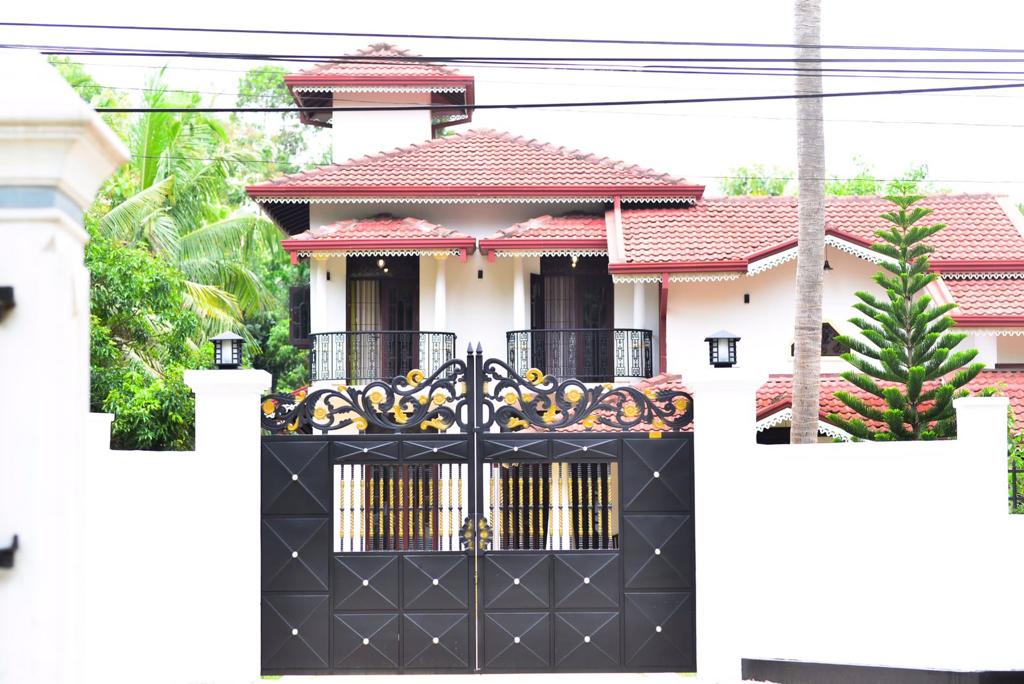 An elegant villa is framed by a decorative black gate with intricate designs. The building features a tiled roof and balconies, surrounded by vibrant greenery. The welcoming entrance includes white walls and lanterns, enhancing the appeal of the property.