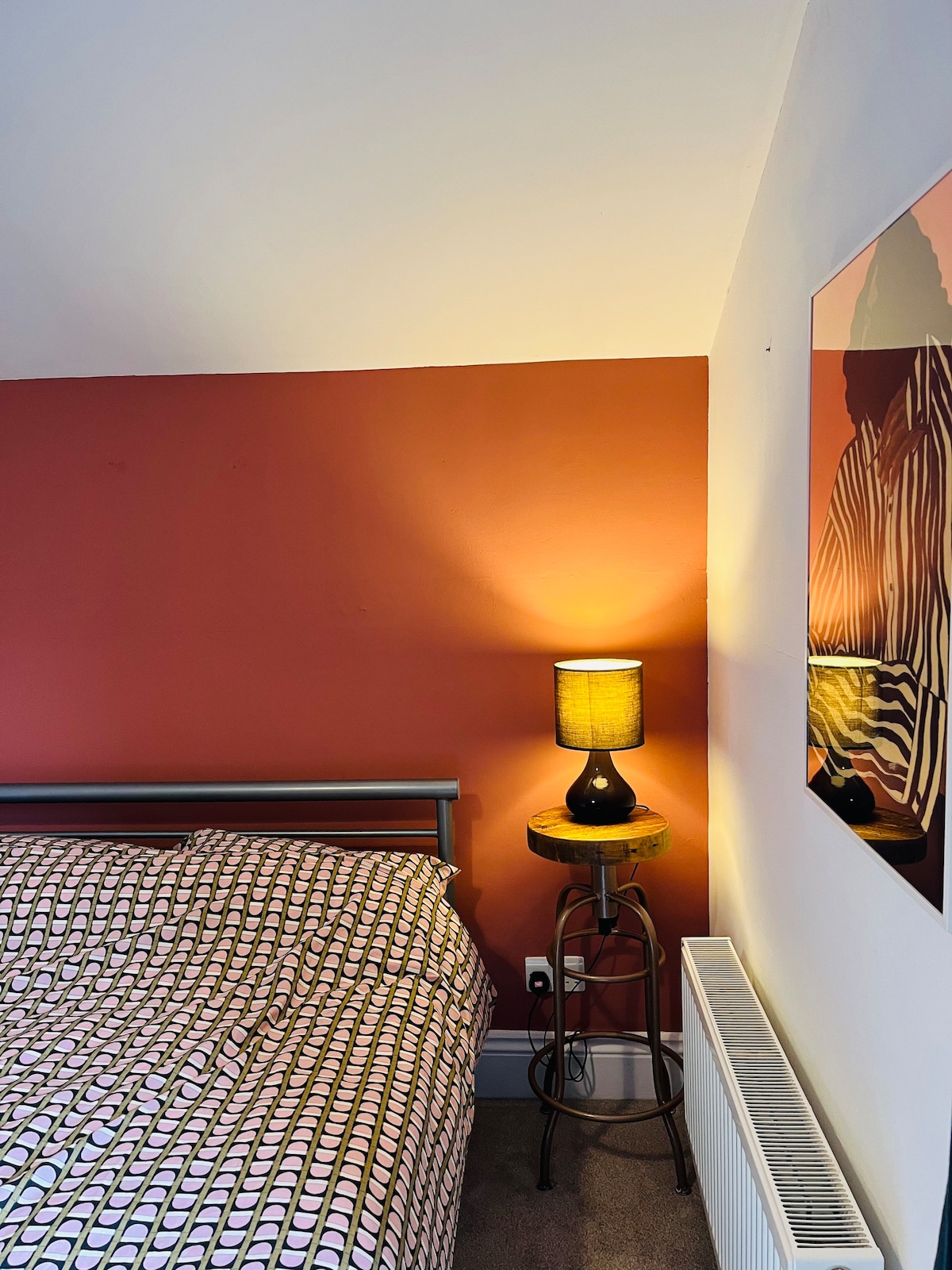 A cozy bedroom corner showcases a spacious bed adorned with patterned bedding. A warm table lamp sits on a stool beside the bed, casting a soft glow. A mirror reflects the room's light and adds depth to the space.