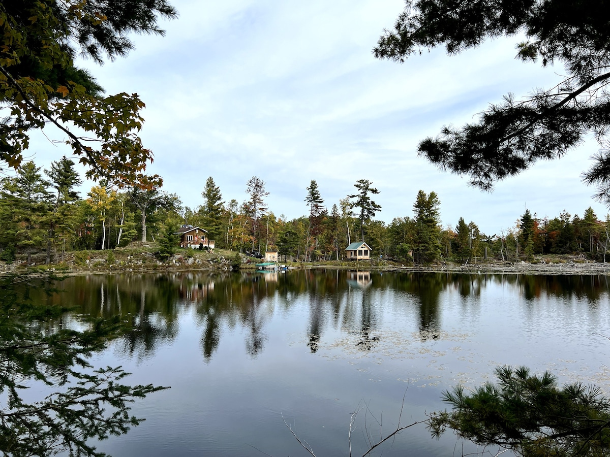 A serene lake is framed by lush trees and scattered cottages. The mirrored surface reflects the surrounding nature and sky, creating a calm atmosphere. Soft neutral tones dominate the scene, with subtle hints of color from the foliage, indicating a peaceful backcountry setting.