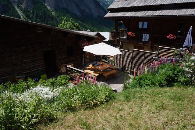 An outdoor terrace is visible, featuring a large table covered by a white umbrella. Surrounding greenery and colorful flowers enhance the space, while wooden buildings rise in the background against a mountainous backdrop.