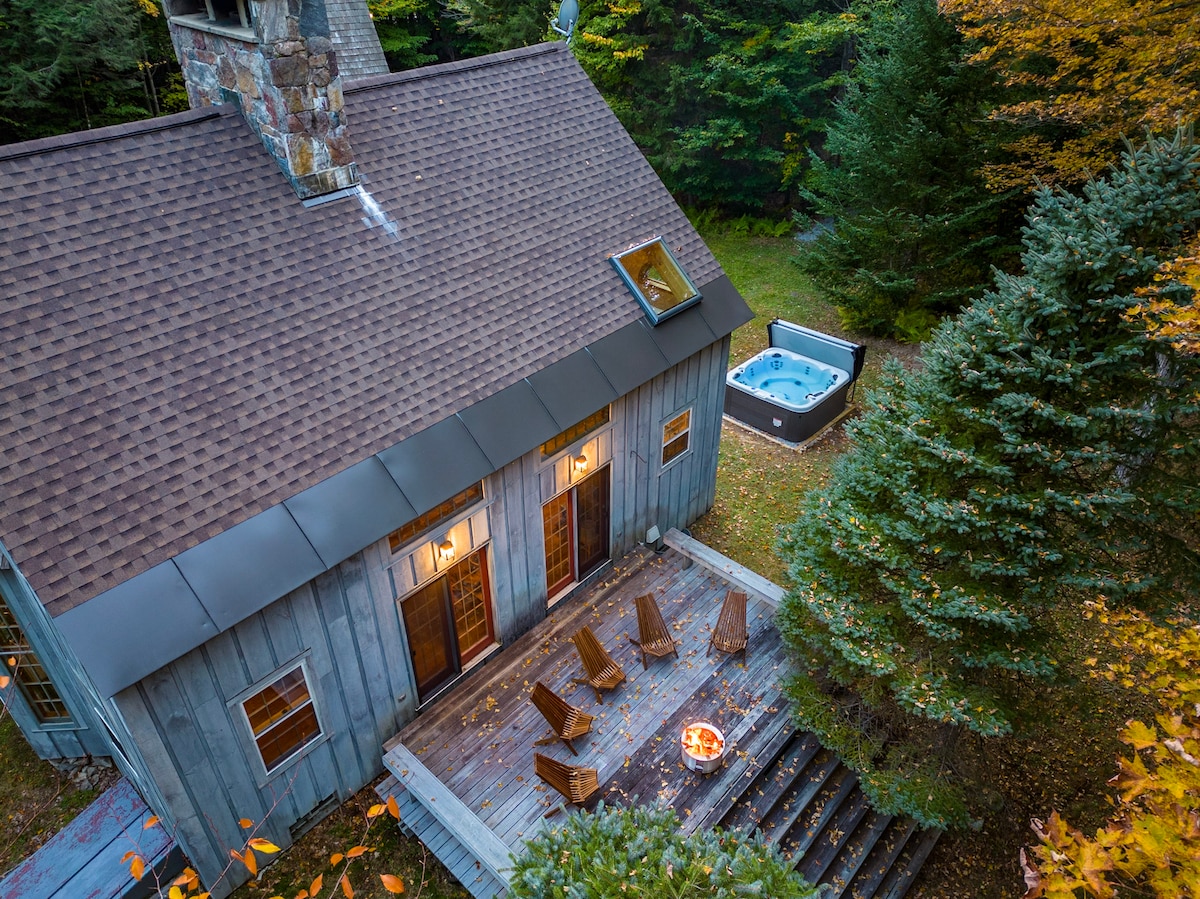 An aerial view showcases a cabin with a wooden exterior and a sloped roof. A spacious deck is furnished with several chairs surrounding a fire pit. Beside the deck, a hot tub is nestled among the trees, highlighting the cabin's serene outdoor setting.