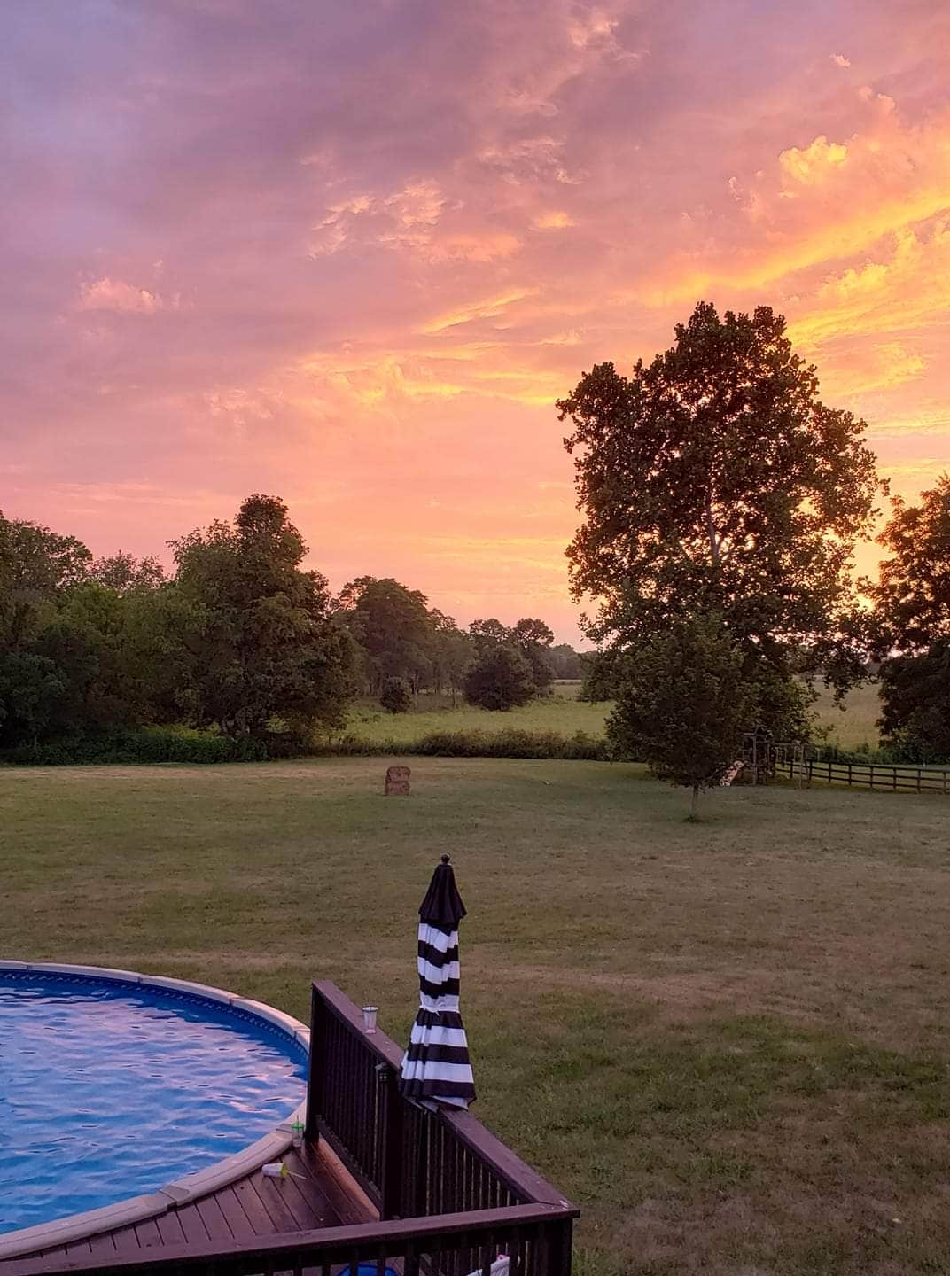 The image features a round above-ground pool surrounded by an expansive, grassy area. A large tree stands to the right, with rolling hills visible in the background and a colorful sunset sky painted in shades of orange and pink.